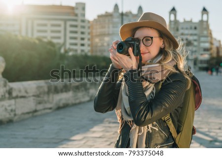 Sunny day, autumn. Young woman tourist, photographer, hipster girl dressed in hat and eyeglasses, stands on city street and takes photo. Vacation, travel, adventure, sightseeing. Blurred background.