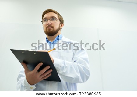 Low angle  portrait of young bearded doctor wearing glasses standing against white wall writing on clipboard, copy space