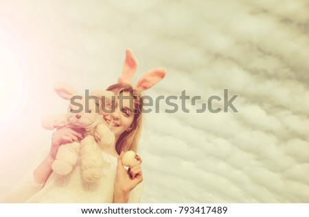 happy easter, traditional spring holiday celebration, girl in pink bunny ears with egg and rabbit toy, has long blonde hair and smiling adorable face outdoor on cloudy sky background, copy space