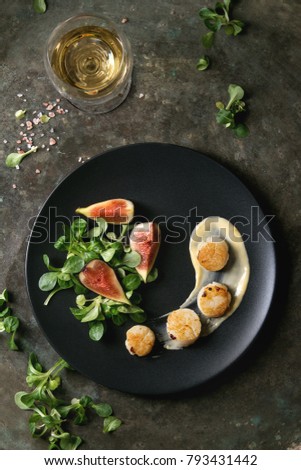 Fried scallops with lemon, figs, sauce and green salad served on black plate with glass of white wine over old dark metal background. Top view, space. Plating, fine dining Royalty-Free Stock Photo #793431442