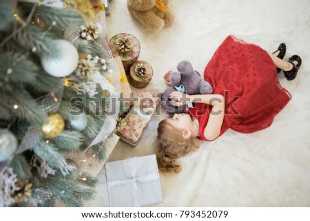 A little girl is lying under a Christmas tree with a teddy bear next to boxes with gifts