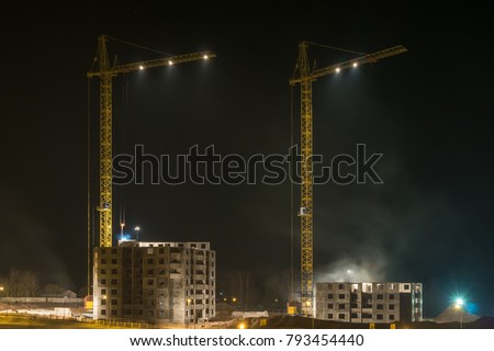 Tower cranes and unfinished multi-storey high buildings under construction at night on illuminated building site
