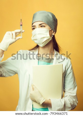 Beautiful young doctor in mask and medical uniform holding syringe with liquid and folder on orange background