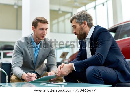 Portrait of handsome young man signing purchase contract buying new car in luxury showroom, sitting at table with mature salesman