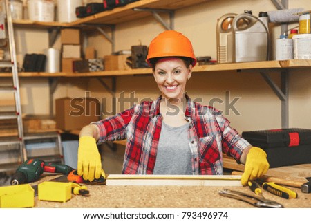 Young woman in plaid shirt, gray shirt, yellow gloves, protective helmet measuring length of piece of wood with tape measure, working in carpentry workshop at wooden table place with different tools