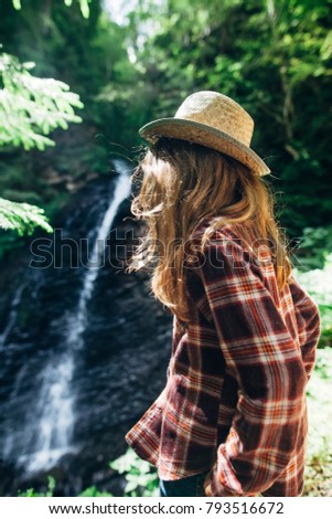 Back view of young girl in shirt and straw hat from afar admires waterfall. Caucasian young lady looks at falling water, photo on fresh air. Portrait of youth adolescent in wild nature