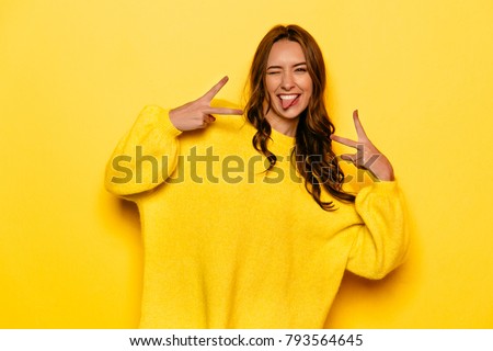 Smiling pretty girl with curly hair winking, showing a tongue, peace gesture. On yellow background.