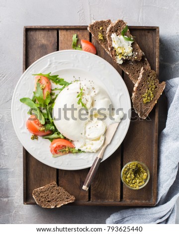 Sliced Italian cheese Burrata with a vintage knife, fresh arugula, tomatoes, olive oil, pesto sauce and whole grain bread toasts on a wooden box and a concrete light background.  Mmm, yummy.)
Flat lay
