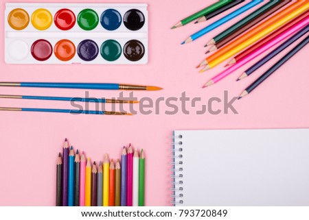Multicolored set of pencils, watercolor and notepad laying on pastel pink background top view. Creative set for hobby