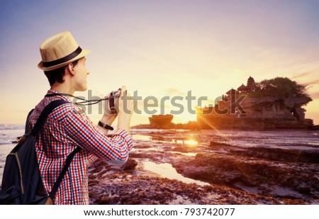 Travel and photography. Young man with camera taking picture of beautiful balinese landscape. Ancient hinduism temple Tanah lot on the rock against sunset sky. Bali Island, Indonesia.