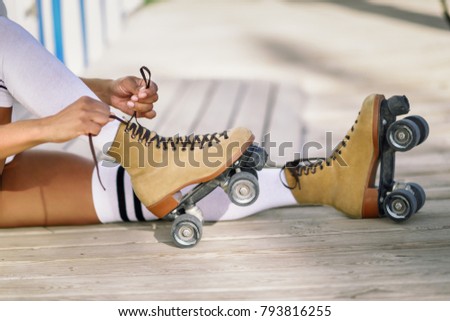 Close-up of black girl sitting on wooden floor puts on skates outdoors. Unrecognizable woman. 