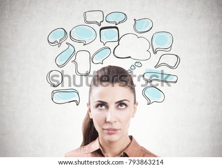 Portrait of an attractive young brunette woman wearing a brown blouse and looking upwards at white and blue speech bubbles drawn on a concrete wall. Mock up