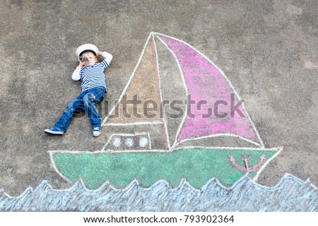Little kid boy as pirate on ship or sailingboat picture painting with colorful chalks on asphalt. Creative leisure for children outdoors in summer. Child with captain hat and binoculars