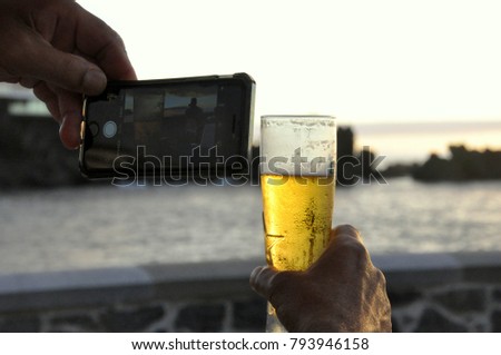 man taking picture with smart phone of glass of beer during sunset