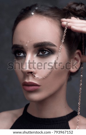 Girl with cool fashionable make-up posing in studio,matte lips and nails,cate eyes,smokey gold eyes, beautiful make up and piercing chain from nose over purple dark background with magic glow.