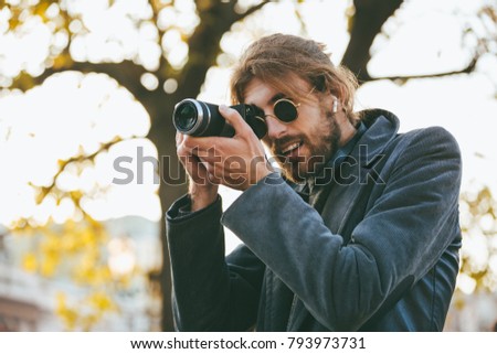 Portrait of an attractive bearded man wearing sunglasses and coat standing on a city street and taking a picture with a camera