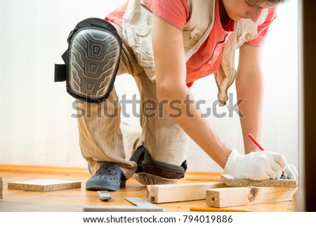 Carpenter young guy in gloves and knee pads marks wooden bars Royalty-Free Stock Photo #794019886