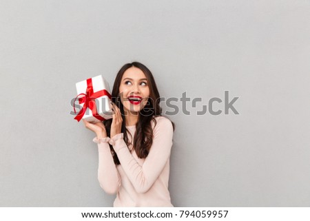 Image of lucky woman holding bithday present with red bow, being happy to win prize being isolated posing over gray background
