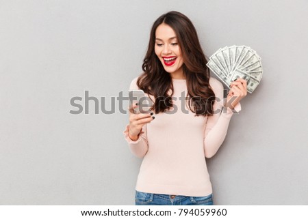 Portrait of attractive brunette female 30s winning lots of money dollar currency using her smartphone, being joyful over gray wall