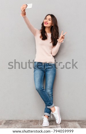 Full-length image of joyful woman smiling with white teeth and making selfie photographing herself, showing peace sign over gray background