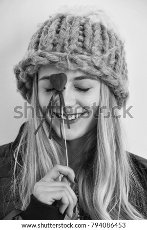 Pretty smiling girl or cute young woman with blond hair and adorable happy face in fashionable grey hat has big blue eyes holds red valentines day heart on stick on white background