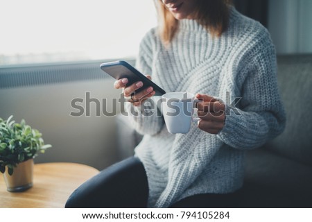 Happy smiling hipster girl using smartphone device while chilling at home wearing cozy knitted sweater, attractive young woman chatting with friends at social network while sitting on sofa at home