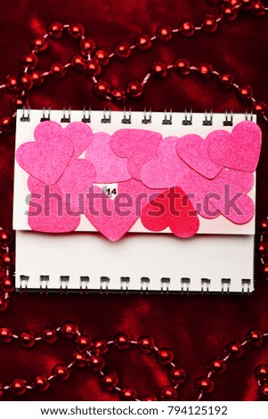 Pink paper hearts covering flip calendar page. Date of Valentines day highlighted. Calendar on red velvet background surrounded by red beads. Love and holiday concept.