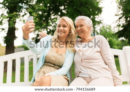 family, technology and people concept - happy smiling young daughter and senior mother with smartphone sitting on park bench and taking selfie