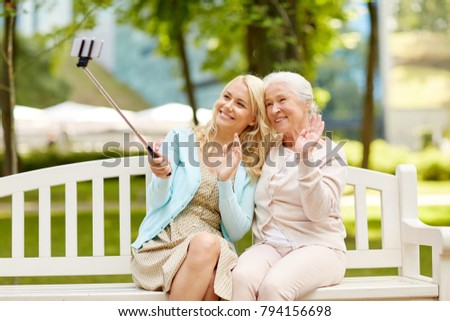 family, technology and people concept - happy smiling young daughter and senior mother sitting on park bench and taking picture with smartphone selfie stick