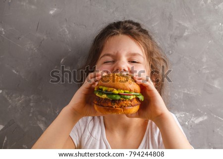 The little girl is eating a healthy baked sweet potato burger with a whole grains bun, guacamole, vegan mayonnaise and vegetables. Child vegan concept