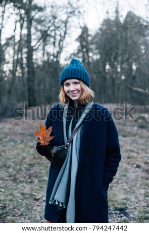 Pretty and cute beautiful red head girl with freckles, in big blue hat, with scarf and coat, stands in middle of forest, picks up fallen leaves and smiles. vertical photo mobile adapted