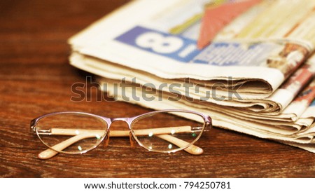 Glasses and pile of newspapers folded and stacked at the background. Selective focus on glasses, blurred pages. Horizontal surface wooden table. Side view