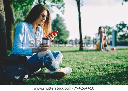Brunette student browsing photos in friends profile on telephone sitting in park with coffee.Female blogger in sunglasses searching music on internet web site for downloading near publisity area