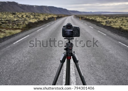 Black camera on the tripod on the roadway with orange roadside pillars between the green fields and mountains on the background of the sea and cloudy sky in Iceland. Closeup. Horizontal.