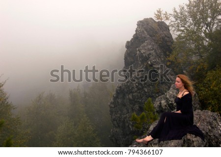 the redhead bare-footed woman in a black dress sits on a rock above the forest against the background of the approaching wall of fog

