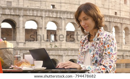 Young woman writing and working on her laptop computer sitting at the table outside in a bar in front of the Colosseum in Rome. Elegant beautiful dress and colorful shopping bags.