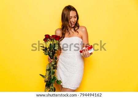 Charming girl looking at gift box, holding a bouquet of red roses received from her boyfriend in St. Valentine's day. Dressed in white dress, curly hair.