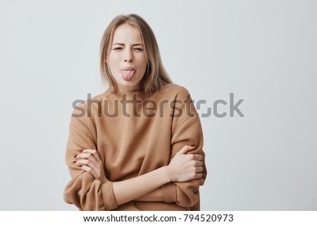 Body language. Naughty stressed beautiful woman with straight fair hair posing against studio wall, frowning her face, keeping arms folded and sticking out tongue, demonstrating disgust to something Royalty-Free Stock Photo #794520973