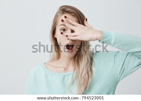 Astonished blonde female keeps mouth widely opened, looks at camera through fingers, remembers some importnat duty which she must do immediately, or is scared of seeing something unpleasant.