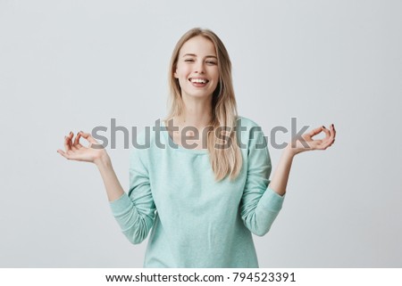 People, yoga and healthy lifestyle. Caucasian young blonde woman keeping eyes closed while meditating indoors, practicing piece of mind, keeping fingers in mudra gesture, smiling broadly
