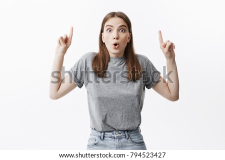 Copy space for advertisement. Attractive young caucasian woman with dark hair and popping eyes looking in camera with surprised look, pointing upside with index fingers on hands.