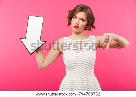 Portrait of a disapointed girl dressed in dress pointing down with an arrow isolated over pink background