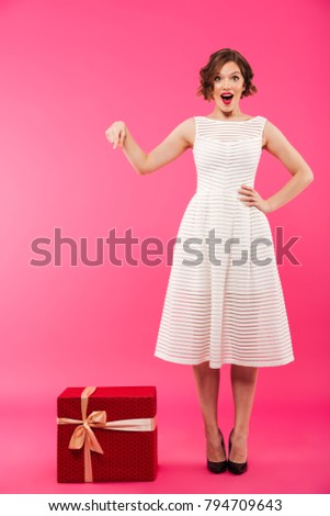 Full length portrait of a lovely girl dressed in dress pointing finger at a gift box while standing isolated over pink background