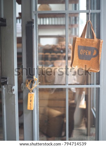 A Metal Grey Door with a Wooden little signage hang on the handle. Also a lovely little brown rough bag with a message 'OPEN' hang at the front of the Café / Coffee shop in the morning 