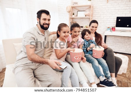 Dad, mom, two girls and a boy watch movies on TV. They are sitting on the couch in their apartment. They eat popcorn. They are a happy family.