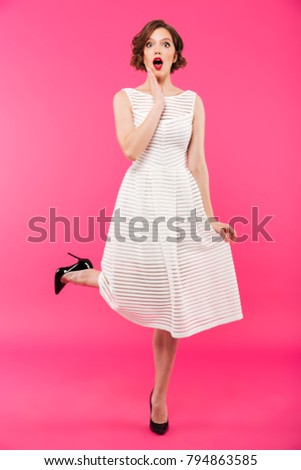Full length portrait of a shocked girl dressed in dress posing while standing isolated over pink background
