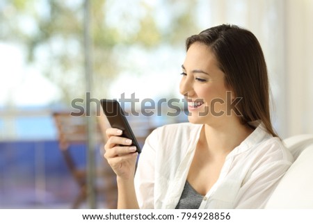 Happy woman using a smart phone sitting on a couch in an apartment with a window and the sea in the background