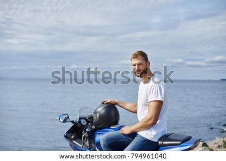 Attractive bearded young motorcyclist enjoying seaview while traveling on his motorcycle along seashore, posing against blue sky and sea background with copy space for your advertising content