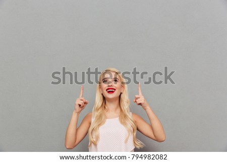 Portrait of a happy girl standing and pointing two fingers up at copy space isolated over gray background