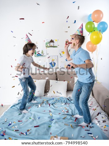 Jolly schoolboys jumping on bed excited by confetti flying around, showing great joy. Balloons on background